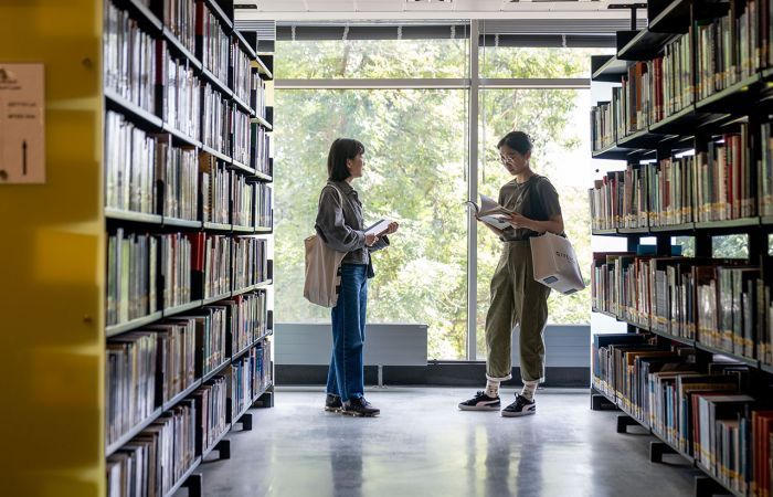 Two students stand in a library, one reads a book