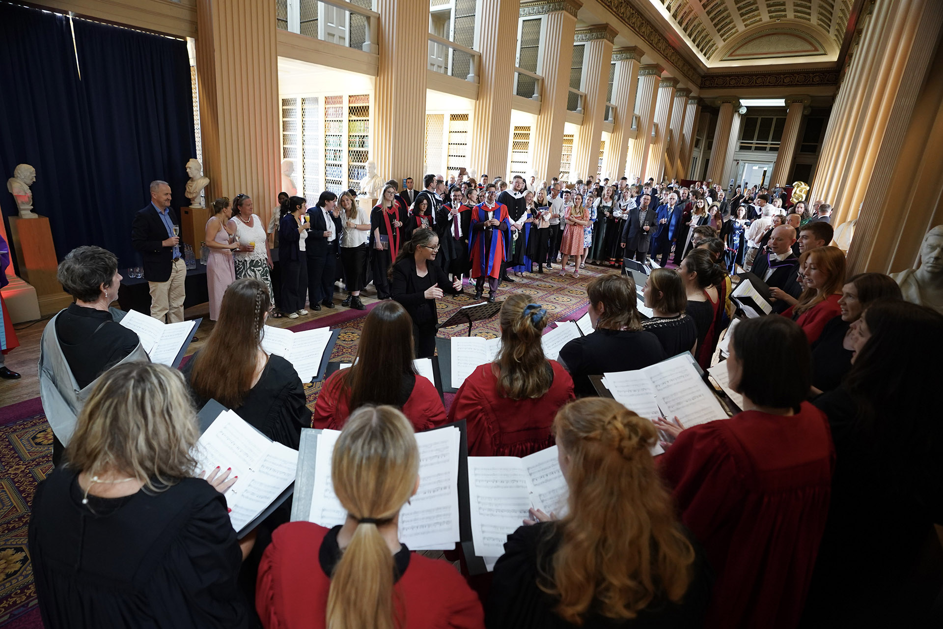 A room of people wearing gowns watch a choir sing from sheet music