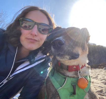 Picture of a white person with long hair wearing a blue jacket and green sunglasses. There is a brown, black and white dog wearing a green harnesses and red collar. They are on the beaching and the sun is shining brightly in the background.