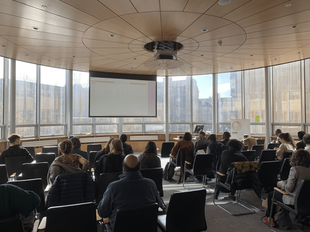 Photo taken from the back of the conference room, with people seated and looking at a projector screen
