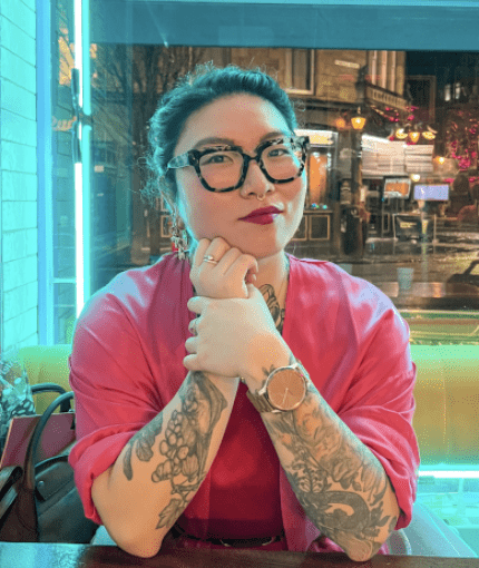 a tattooed Korean woman with glasses sitting at a table backlit with blue neon lights