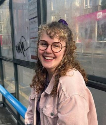 A picture of a woman with curly hair, she is smiling and wearing a pink jacket.