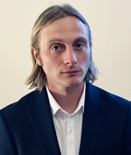 Man in his thirties with long blond hair, blue eyes in a blue suit jacket ad white shirt.
