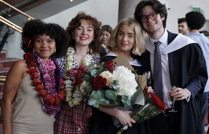 Four graduates, wearing robes and carrying flowers, smile at the camera