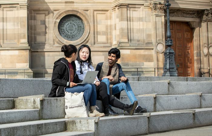 Three students sit on steps outside with a laptop