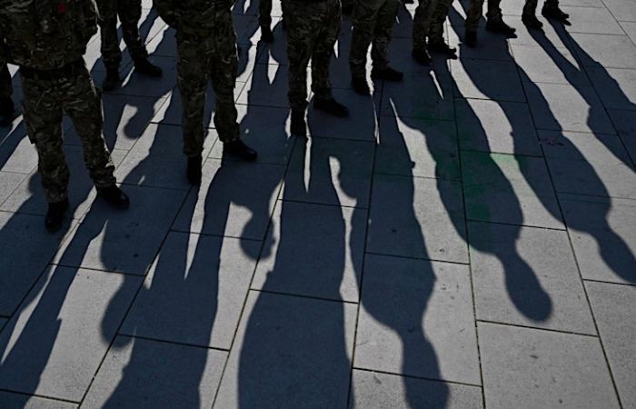 A group of people in army uniforms stand on concrete, there are long shadows on the ground.