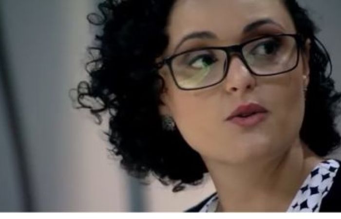 A close up of the face a mixed-race, Brazilian woman, earing dark-rimmed glasses and dark red lipstick. She has chin-level, curly hair, and is in the middle of speaking, as if in an interview.