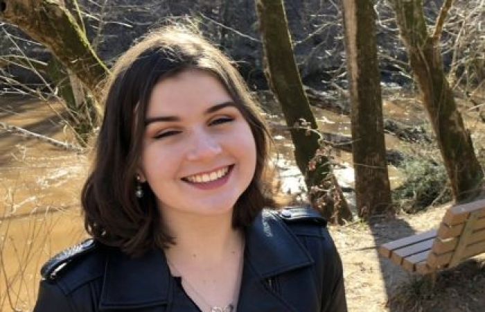 Photo of a smiling person in a leather coat, standing in front of trees.