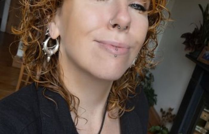 This is a picture of me shoulder-length, curly, slightly wet, ginger-coloured hair. I am smiling and looking at the camera. Here, I have winged eyeliner and multiple facial piercings, including a septum ring, nose and a labret lip piercing. I am wearing large silver hoop earrings, a textured black shirt, and a bold, triangular silver pendant necklace on a black cord. The background shows a warmly lit room with wooden floorboards, indoor plants, and a tiled fireplace decorated with candles and framed photos.