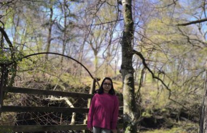 Image of Nabanita wearing a pink jumper, a jeans, and a spectacle. She is standing by a stream of water, with the blue sky and few trees visible around her. 