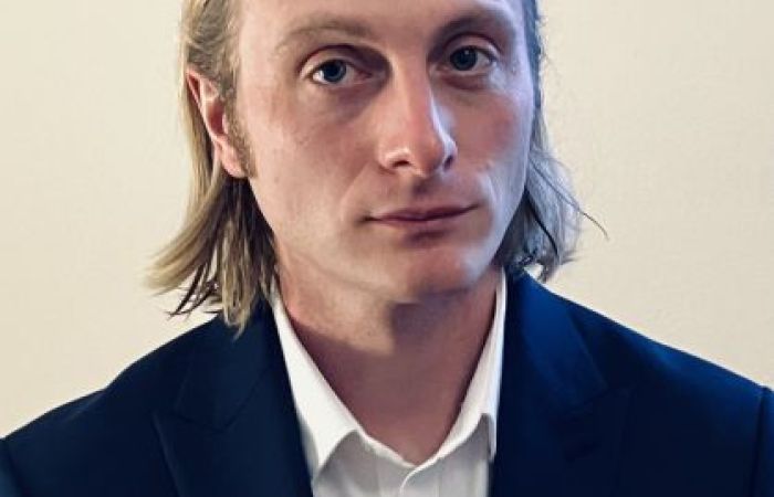 Man in his thirties with long blond hair, blue eyes in a blue suit jacket ad white shirt.